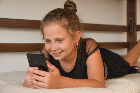 Technology, free time, communication concept. Cheerful teengaer dressed in nightwear, uses mobile phone for online chatting, networking, recieves text message, lies on bed