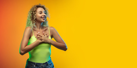 Excited, shy. Caucasian young woman's portrait on gradient studio background in neon. Beautiful female curly model in casual style. Concept of human emotions, facial expression, youth, sales, ad