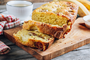 Loaf of banana bread on a wooden cutting board with cup of tea on rustic wooden table.