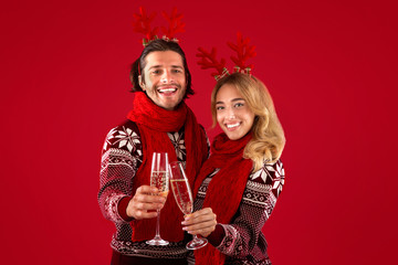 Millennial couple in sweaters and deer horns celebrating Christmas or New Year with champagne, red background