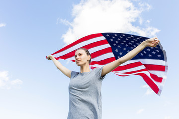 Young woman with a serious expression face holding an American flag on protest. Demonstration. USA flag against the sky