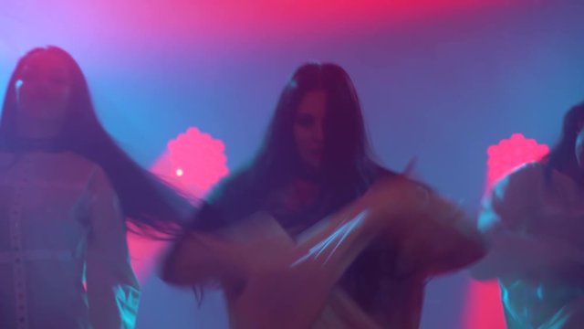 Silhouettes of a three charming womans in white shirt are dancing on the stage. Shot in a dark studio with smoke and neon lighting. Red neon light effects. Dance show.