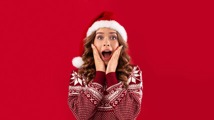 Christmas celebration. Excited young woman in Santa hat and warm winter sweater over red background