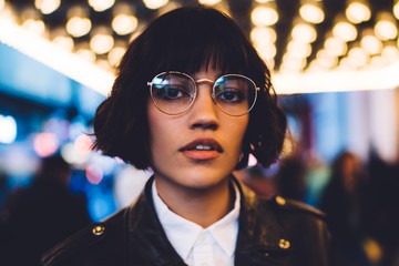 Close up portrait of female model dressed in trendy clothing have evening sightseeing around illuminated street in megalopolis, stylish hipster girl in glasses looking at camera during travel