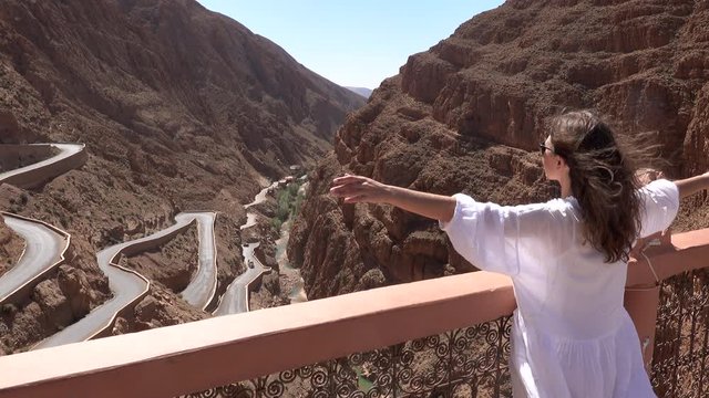 Tourist woman in white dress raising hands up and enjoy view of windy mountain road in the Dades Gorge, Gorges Du Dades, Morocco