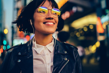 Cheerful woman teenager in glasses with neon lights spending evening in megalopolis and relaxing with positive music,youthful hipster girl in electronic headphones listening audio playlist in New York