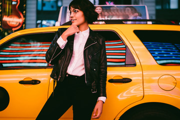 Caucasian trendy dressed woman standing on urban setting with cab taxi on background in night city, gorgeous hipster girl in eyewear looking away during getaway travel for recreating in New York