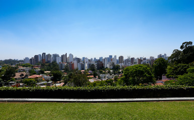 Quintal de casa com grama aparada e a cidade de São Paulo ao fundo, em dia ensolarado com céu sem...