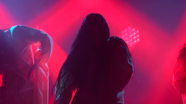 Silhouettes of a three charming womans in white shirt are dancing on the stage. Shot in a dark studio with smoke and neon lighting. Red neon light effects. Dance show.