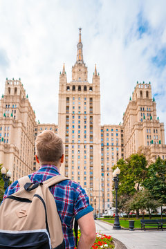 A young man with a backpack stands looking at the Old high rise in the center of Moscow