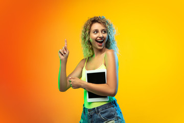 Using tablet. Caucasian young woman's portrait isolated on gradient studio background in neon. Beautiful female curly model in casual style. Concept of human emotions, facial expression, youth, sales