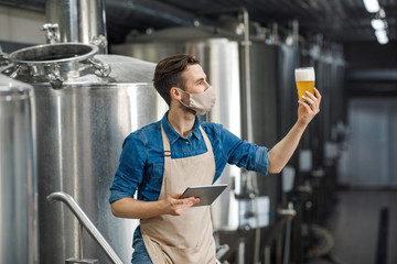 Beer quality control. Man with digital tablet looking at glass of beer on factory