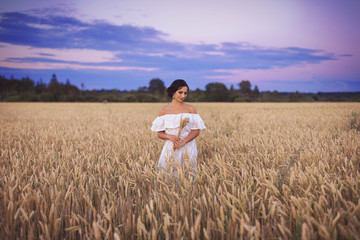 Portrait of a young beautiful brunette girl in a white summer dress standing in a field with wheat ears on the background of the sunset sky