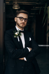 Handsome young groom with beard and glasses dressed in a classic black suit and white shirt and bow-tie sitting. Male portrait. Stylish man dressed in a black suit