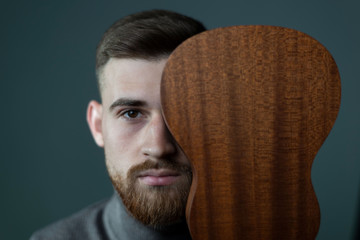 a portrait of a twenty six year old bearded young man covering half of his face with a ukulele. On...