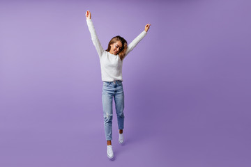 Cheerful student in good mood happily jumping in studio on isolated background. Long-haired brown-haired woman in stylish jeans and white sneakers poses for full-length photo