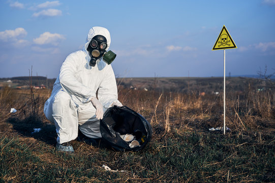 Portrait of ecologist in protective suit and gas mask. Research scientist picking up garbage in grassy field with warning symbol of poisonous substances and danger. Concept of environmental pollution