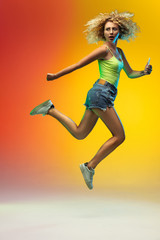 Fototapeta na wymiar Hurrying up. Jumping caucasian young woman's portrait on gradient studio background in neon. Beautiful female curly model in casual style. Concept of human emotions, facial expression, youth, sales