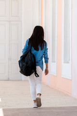 Woman with long brown hair walking with backpack. View from the back. White jeans and blue shirt. Light hall background.