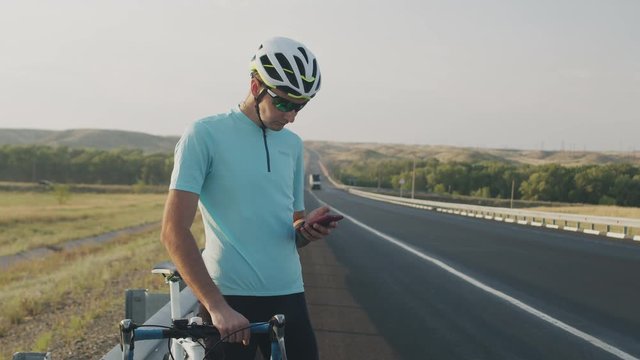 A young male cyclist stands at the curb next to the bike, looking at the phone, flipping through the screen and looking away