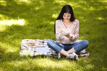 Smiling asian girl making notes in notepad while sitting outdoors in park