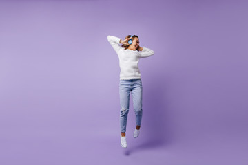 High-spirited lucky female student jumping in studio on isolated background. Full-length shot of girl in fashion white sweater and light blue jeans.