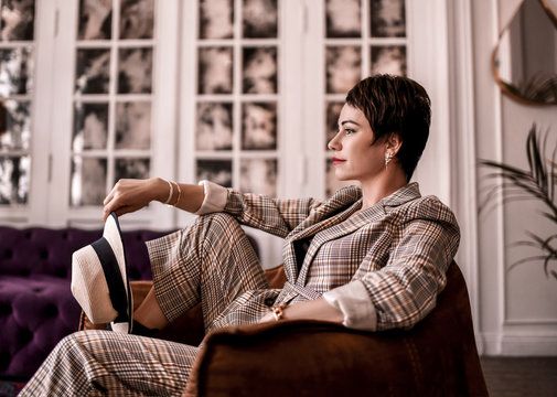 Calm Smiling Brunette Woman In Elegant Business Plaid Pantsuit With Rolled Up Sleeves Sits In Armchair. Side View