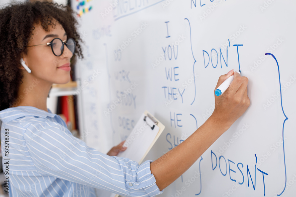 Wall mural Online teacher. African american girl in glasses with wireless headphones writes with marker on blackboard