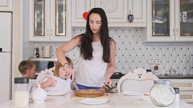 Pretty mother with little son and daughter cutting raspberry pie in kitchen. Happy family of mother and two kids together. 4K, UHD
