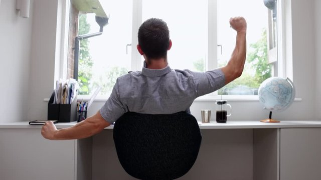 Rear View Of Man Working From Home On Computer  In Home Office Stretching At Desk