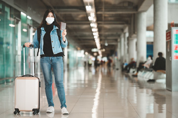 Happy Asian woman tourist wearing face mask holding passport with luggage at airport terminal during coronavirus or covid-19 outbreak . New normal travel at airport