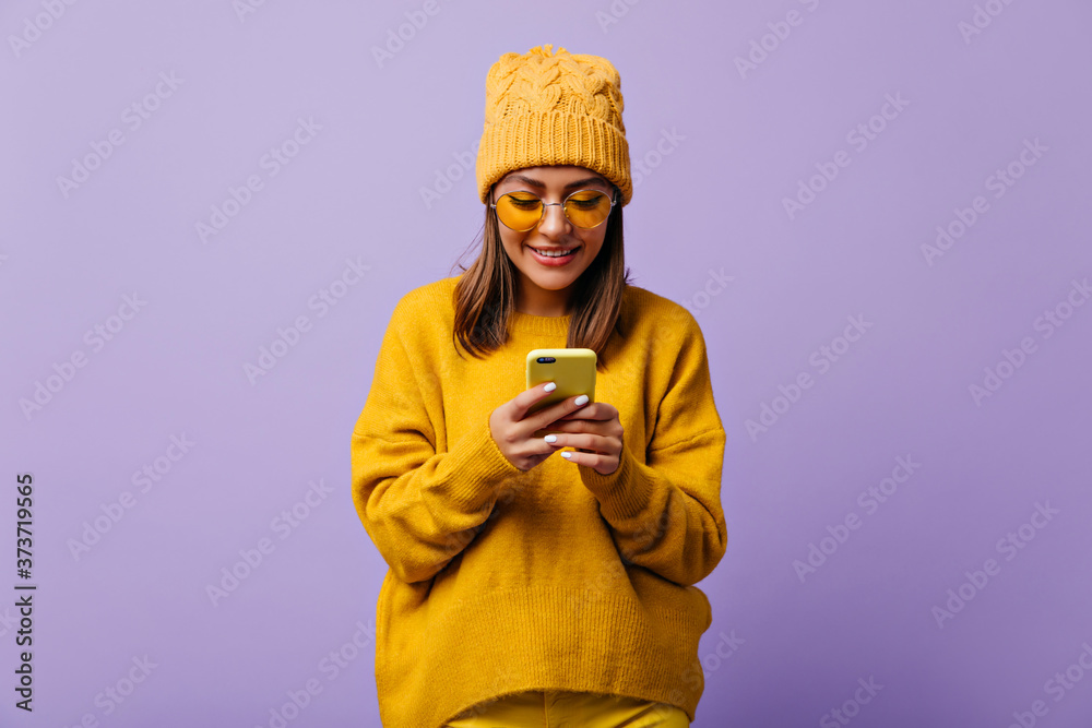 Wall mural Lovely glad woman likes yellow color and wears total yellow outfit. Close up studio snapshot of beautiful girl chatting in her smartphone