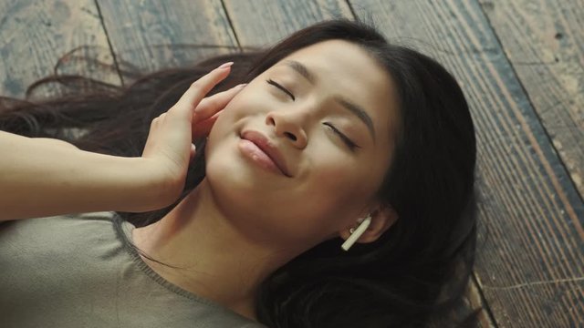 A top close-up view of a happy young asian woman with earbuds is listening the music laying on the floor at home