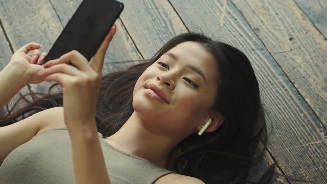 A top close-up view of a smiling young asian woman with earbuds is typing on the phone laying on the floor at home