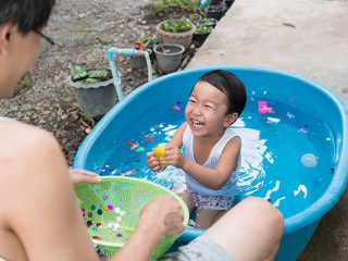 Asian cute child boy laughing while playing water in blue bowl with relaxing face and wet hair in rural nature. Young kid having happy moment in summer. Family activity at home and preschool concept.