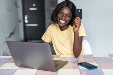 Pretty young african woman using laptop and holding credit card in kitchen