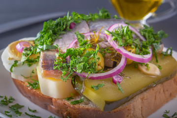Sandwich with herring fillets, onions, pickled cucumber, egg and herbs on a white plate on a dark table. Traditional Scandinavian street food. Close-up