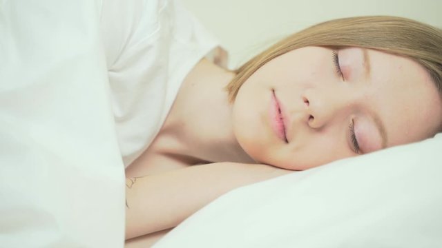close-up portrait of a young woman lying on a bed on a pillow. Student girl sleeps on a white bed with her hands under her head. Blonde with closed eyes resting in the bedroom at home
