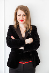 A blonde girl in a black suit stands against a white wall with crossed arms and looks into the camera