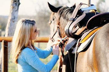 Western lifestyle shows woman putting on saddle to go horseback riding outdoors. - obrazy, fototapety, plakaty