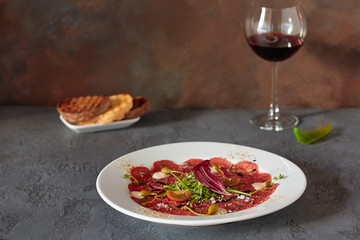 Meat carpaccio in a white plate on a wooden background, with delicious bread and wine in a glass