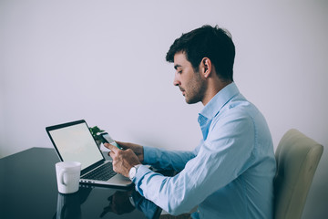 Serious adult executive man using smartphone while sitting at laptop with mug of hot drink