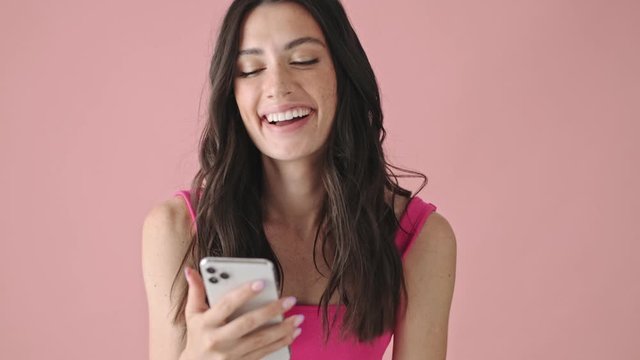 A surprised emotional young woman is typing on her smartphone isolated over a pink background