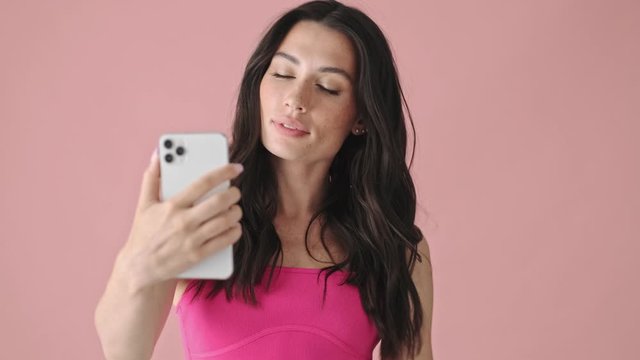 A beautiful young woman is taking selfie photo using her smartphone standing isolated over a pink background