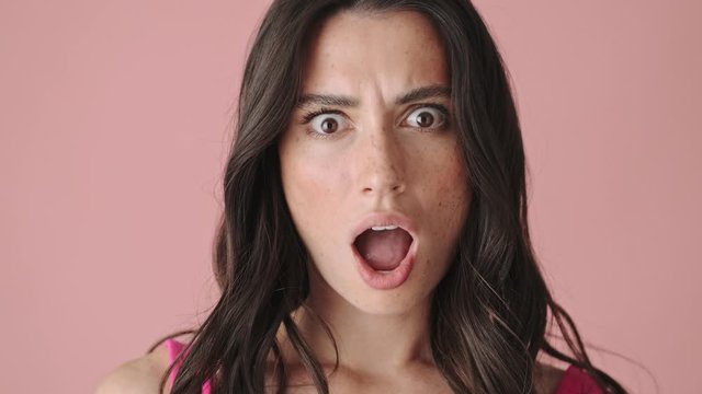 A close-up view of a shocked young woman is looking to the camera isolated over pink background
