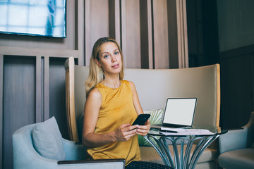 Portrait of blonde caucasian female employee holding mobile phone and looking at camera, confident business woman sending text messages on smartphone app sitting near laptop computer 