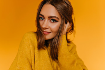 Appealing brown-eyed woman playing with her hair. Stunning white girl in sweater gently smiling on orange background.