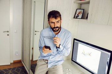 Fototapeta premium Young attractive bearded working dad sitting in his office, thinking and using smart phone.
