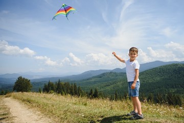 Fototapeta premium Little school age boy running down the slope with kite in the sun. Sunny summer or spring day at sunset. Active outdoor games and leisure. Mountain landscape with hills and coniferous trees.