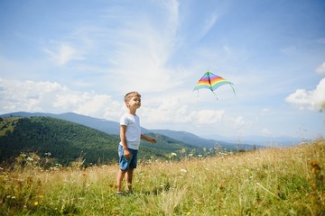 Little boy running on a background of mountains with kite. Sunny summer day. Happy childhood concept.